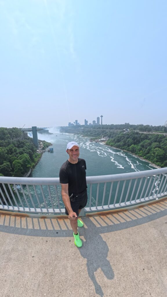 A person is standing on a bridge overlooking a river with waterfalls in the background. The individual is wearing a white cap, black shirt, and bright green shoes, smiling at the camera. The scene includes lush greenery on both sides of the river and a city skyline in the distance under a clear blue sky.