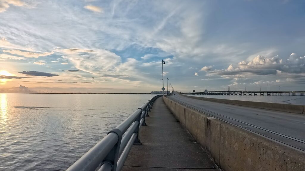 A scenic view of a bridge over a body of water during sunset. The sky is filled with clouds, with the sun setting on the left, casting a warm glow on the water. The bridge has a pedestrian walkway with railings and street lamps. The horizon is visible in the distance, with a mix of blue and orange hues in the sky.