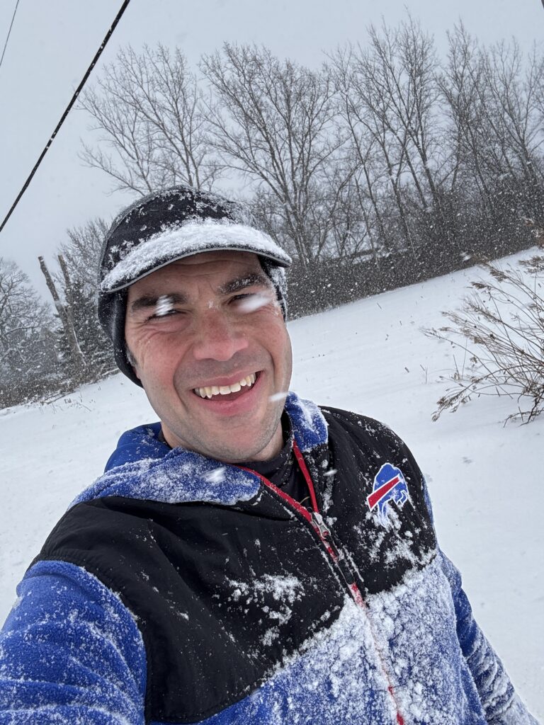 A person is smiling while standing outside in a snowy landscape. They are wearing a black and blue jacket with a sports logo and a black cap, both covered in snow. Snow is falling around them, and leafless trees are visible in the background.
