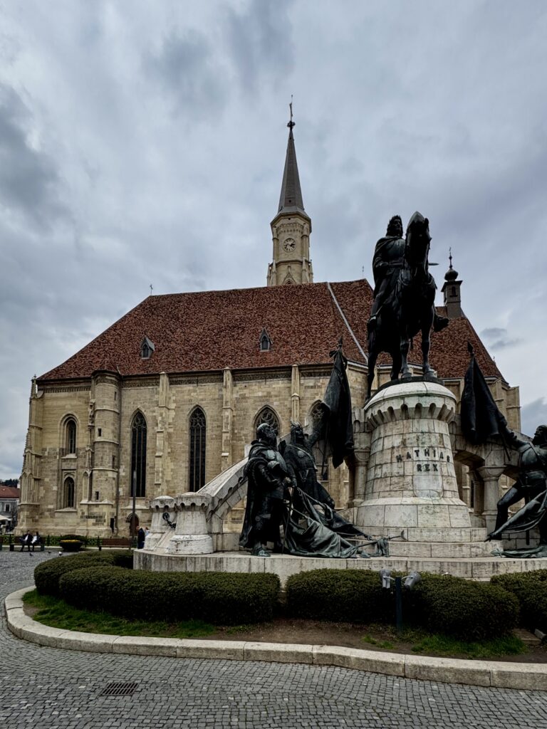 A large Gothic-style church with a tall spire and clock is in the background. In the foreground, there is a statue of a mounted figure on a horse, surrounded by other figures. The base of the statue has the inscription "MATHIAS REX." The scene is set in a paved square with trimmed bushes and a cloudy sky overhead.