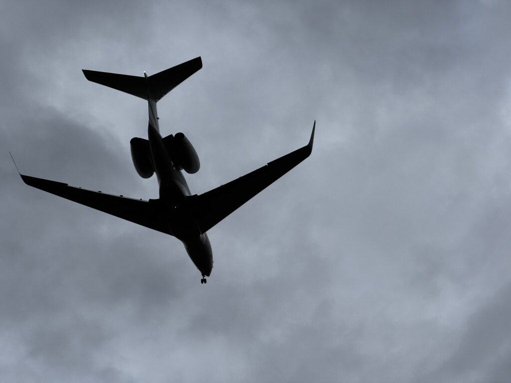 A silhouette of an airplane flying overhead against a cloudy sky. The aircraft is viewed from below, with its wings and tail clearly visible.