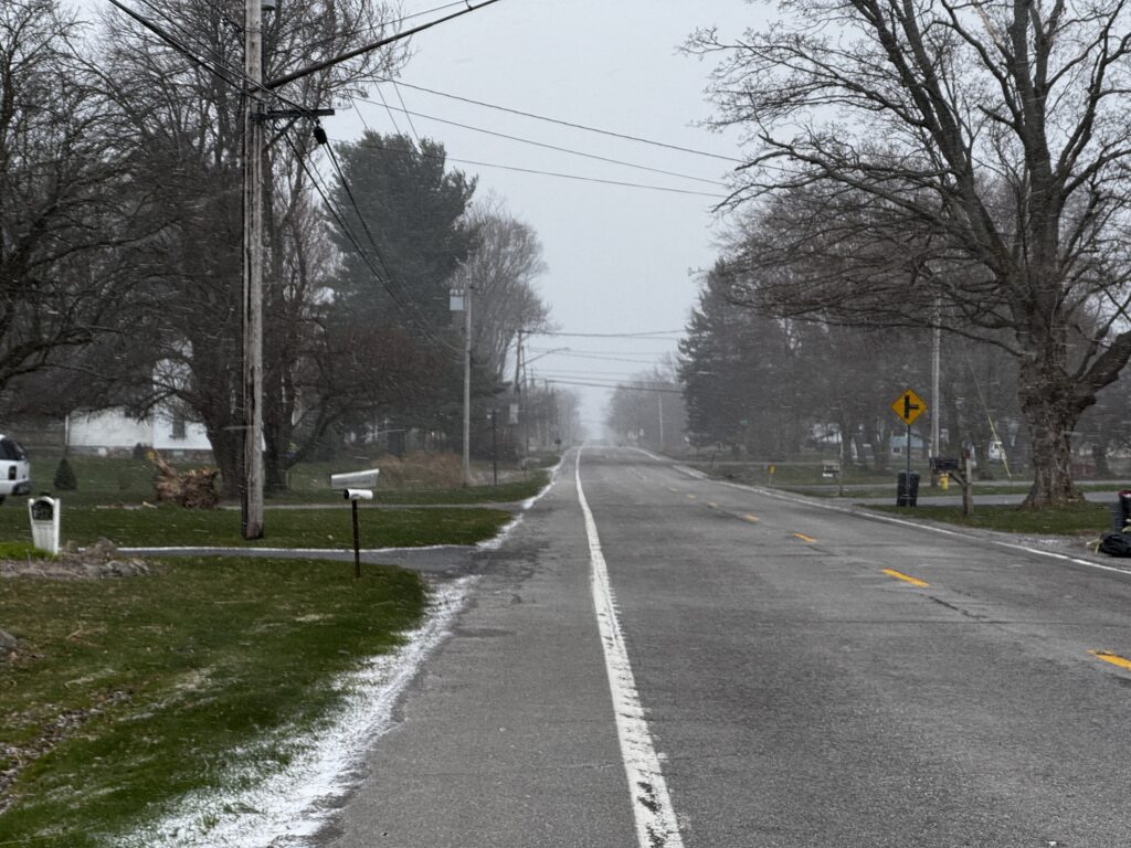 A rural road stretches into the distance, flanked by bare trees and patches of grass. A light dusting of snow covers parts of the ground and road. Power lines run alongside the road, and a yellow road sign indicates a curve ahead. The sky is overcast, suggesting a cold, wintry day.