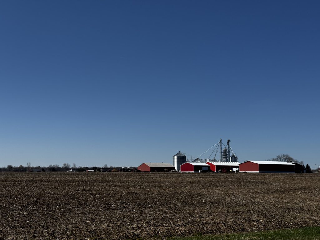 A rural landscape featuring a farm with several red barns and large silos under a clear blue sky. The foreground shows a plowed field, and there are trees and additional farm structures in the distance.