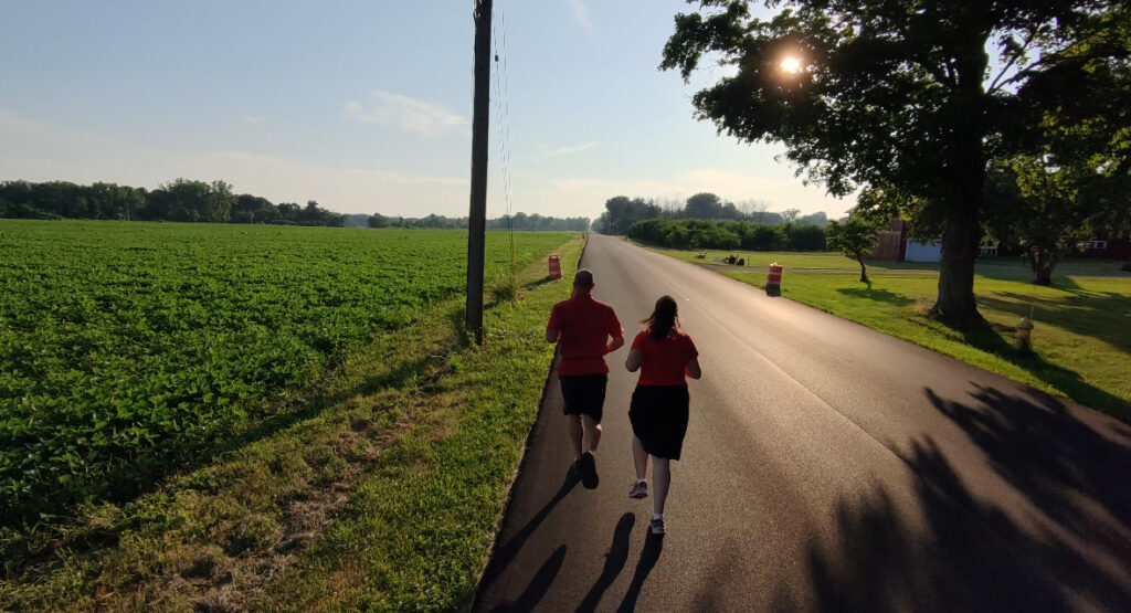 Two people are jogging on a paved road next to a green field under a clear sky. They are wearing red shirts and black shorts. The sun is shining through the trees, casting long shadows on the road. Orange traffic barrels are placed along the roadside.