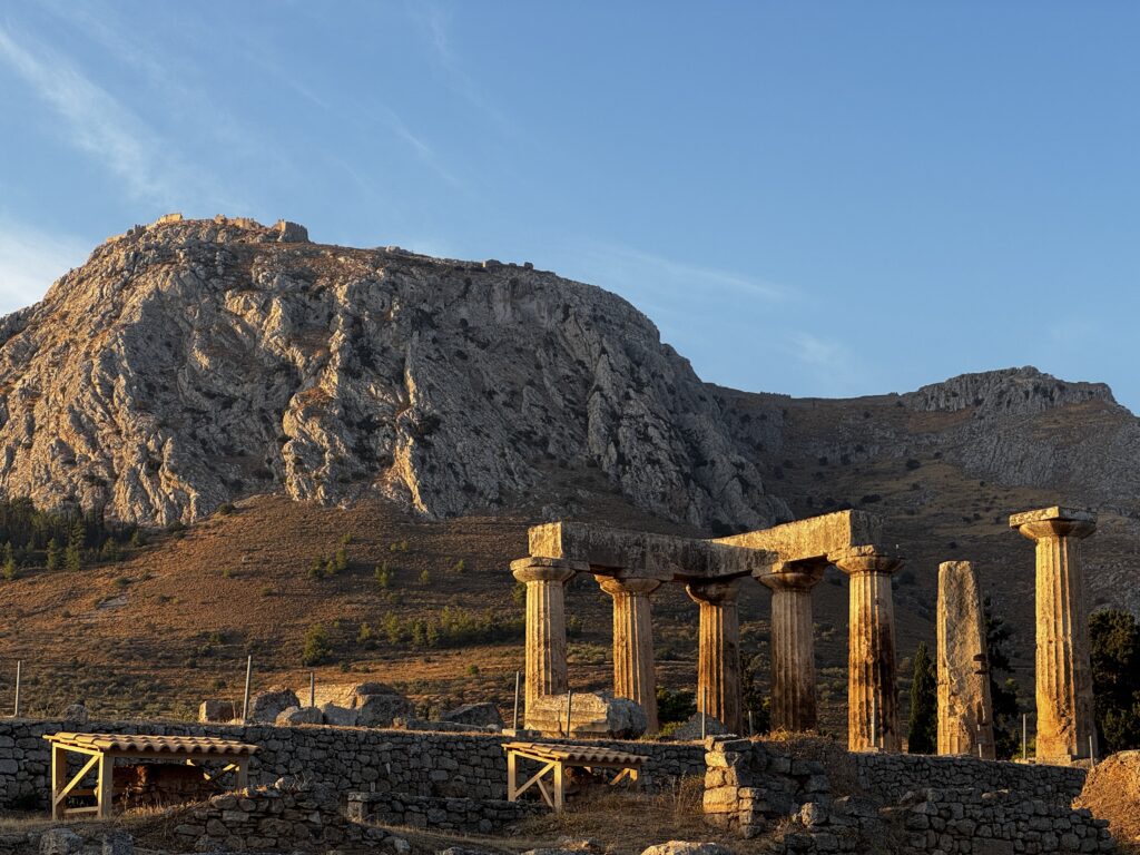 The image shows ancient stone columns and ruins in the foreground, with a large rocky hill or mountain in the background. The scene is bathed in warm sunlight, highlighting the textures of the stone and the landscape. The sky is clear and blue.