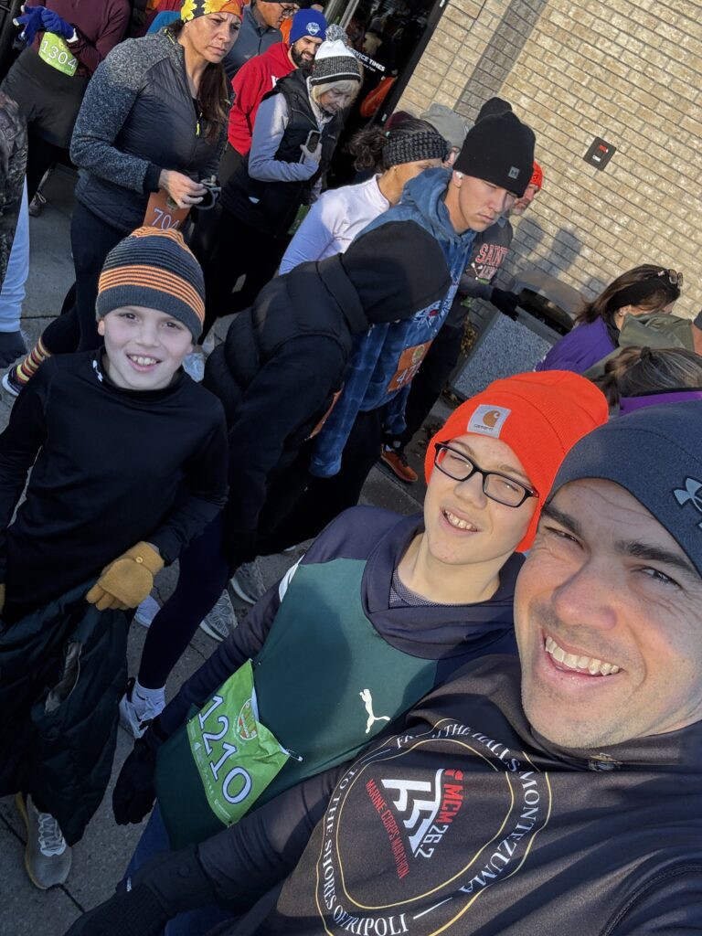 A group of people dressed in winter clothing, including hats and gloves, are gathered outside a building. Some are wearing numbered bibs, suggesting they are participating in a race or event. The atmosphere appears lively and cheerful, with people smiling and interacting with each other.