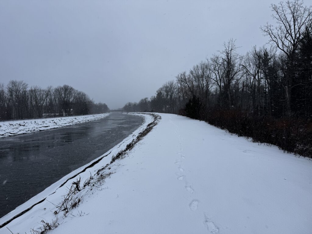 A snowy landscape featuring a river running alongside a snow-covered path. Bare trees line both sides of the river, and footprints are visible on the path. The sky is overcast, and snowflakes are falling gently.