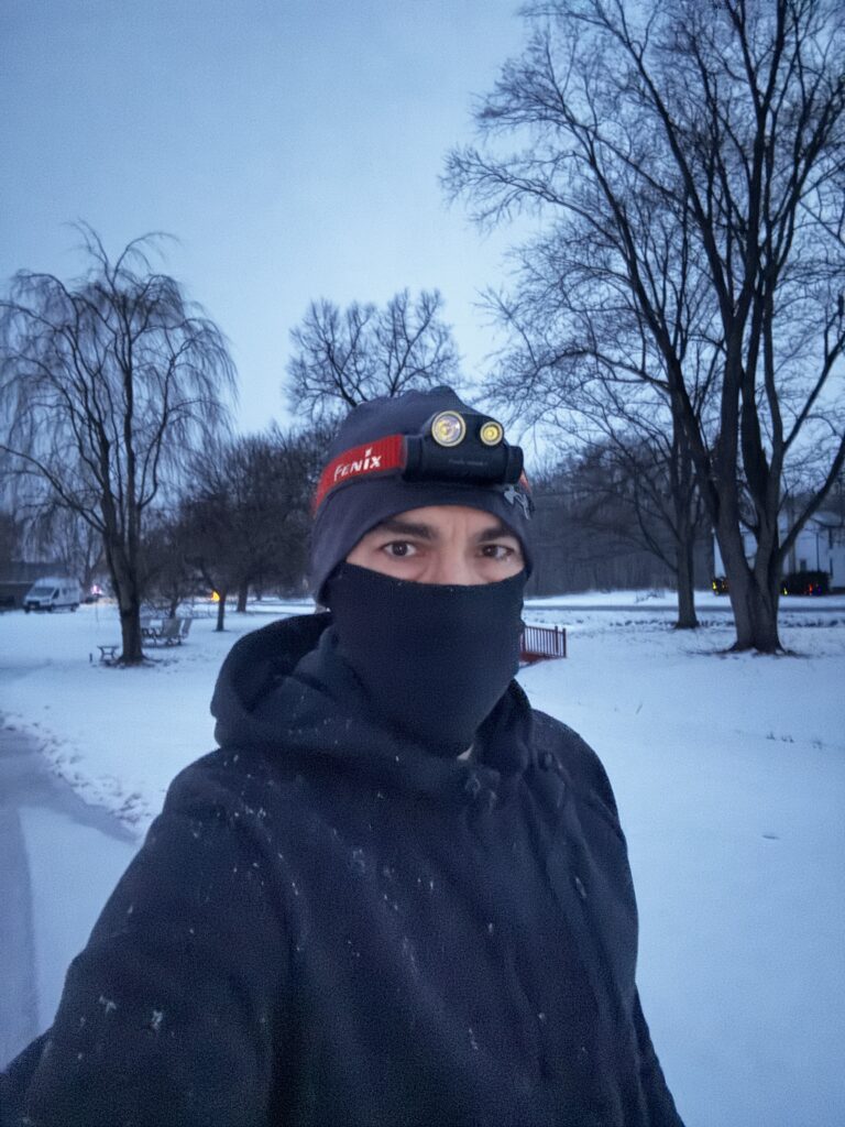 A person is outdoors in a snowy landscape, wearing a black jacket, a black face covering, and a headlamp on a beanie. The background features bare trees and a snow-covered ground, with a cloudy sky overhead.