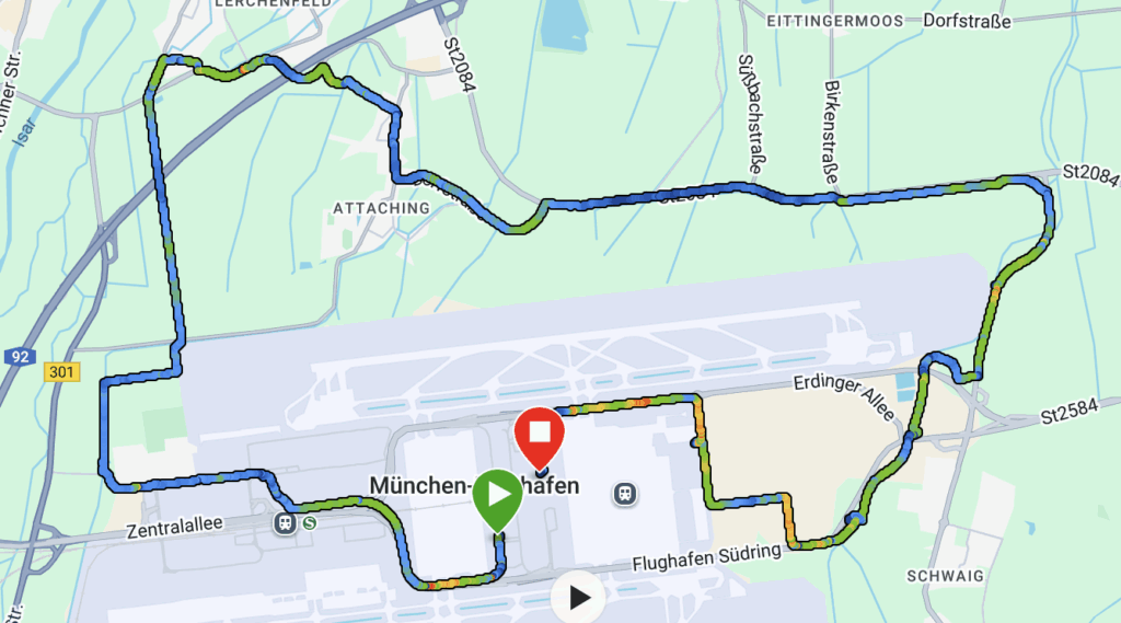 The image is a map showing a route around Munich Airport (München Flughafen). The route is marked with a multicolored line, indicating a path that starts and ends at the airport. The starting point is marked with a green icon, and the endpoint is marked with a red icon. The map includes nearby roads and areas such as Zentralallee, Erdinger Allee, and Schwaig.