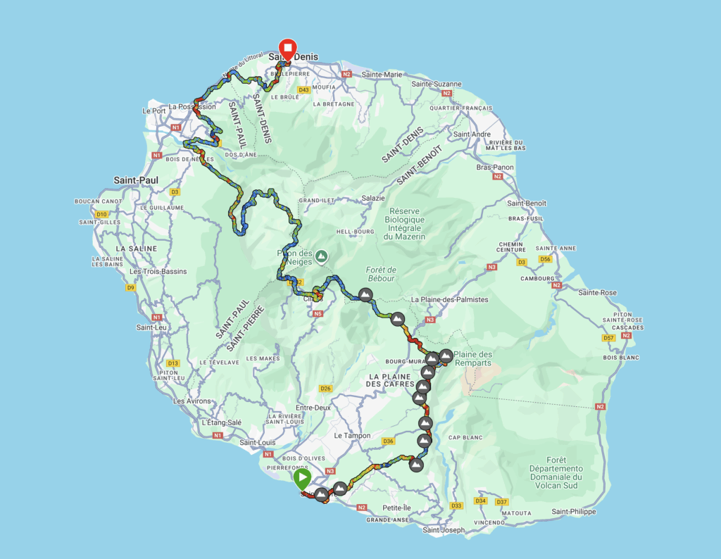 The image is a map of Réunion Island, showing a detailed route for a trail or race. The route is marked with a multicolored line, starting at a green marker in the south near Saint-Pierre and ending at a red marker in the north at Saint-Denis. The path passes through various towns and natural areas, with mountain icons indicating elevation changes. Major roads and towns are labeled, and the map includes geographic features like forests and reserves.