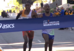Two runners are crossing the finish line of the Los Angeles Marathon. They are both leaning forward, appearing to be in a close race. The finish line tape displays the Asics logo and the words "Los Angeles Marathon." Spectators and event staff are visible in the background.
