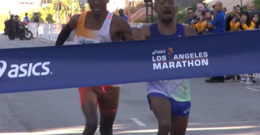 Two runners are crossing the finish line of the Los Angeles Marathon. They are both leaning forward, appearing to be in a close race. The finish line tape displays the Asics logo and the words "Los Angeles Marathon." Spectators and event staff are visible in the background.
