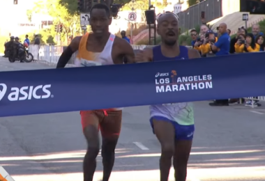 Two runners are crossing the finish line of the Los Angeles Marathon. They are both leaning forward, appearing to be in a close race. The finish line tape displays the Asics logo and the words "Los Angeles Marathon." Spectators and event staff are visible in the background.